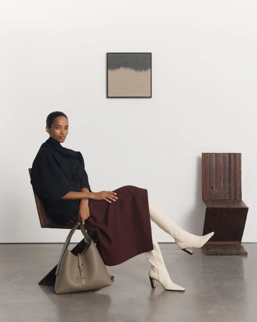 Adult female seated in a modern chair in a light grey room, wearing a black top, dark-red skirt, and white boots; holding a bag. 