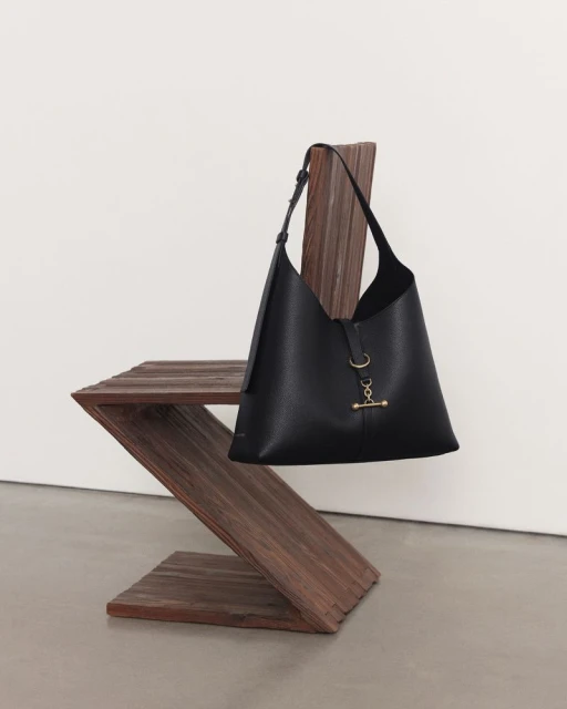Black leather hobo bag hanging on a rustic wooden chair in a minimalist studio setting against a white wall and light gray floor.
