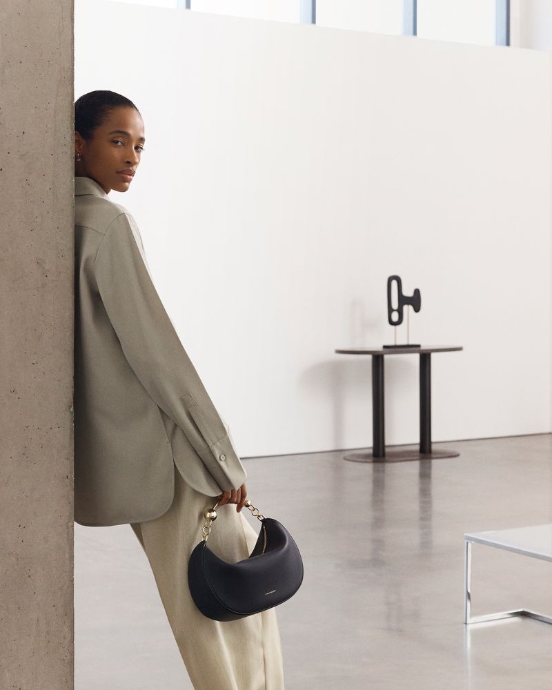 Adult female poses next to a concrete pillar, showcasing a black purse with a gold handle in a minimalist studio setting.