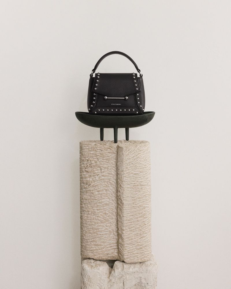 Black leather handbag with studded details on a black display stand set against a clean, bright white backdrop in a studio setting.