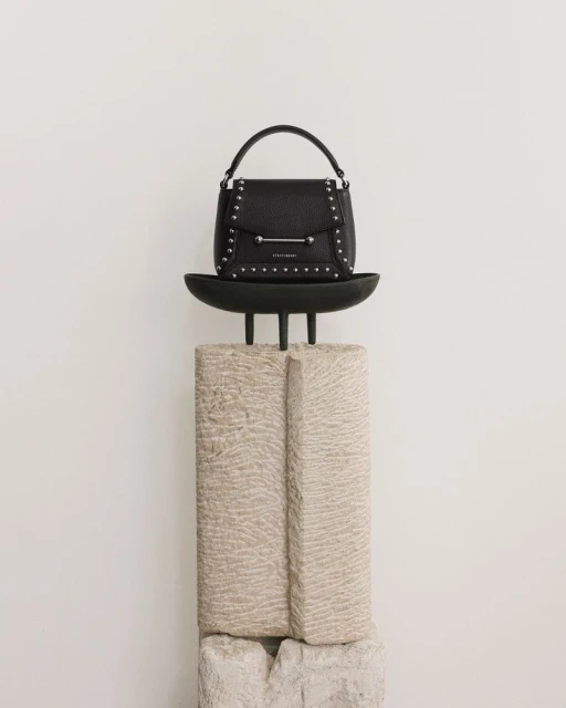 Black leather handbag with studded details on a black display stand set against a clean, bright white backdrop in a studio setting.