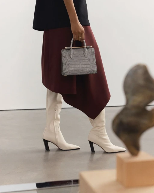 An adult female is shown from the knees down in fashionable white boots and holding a gray handbag with a burgundy skirt.