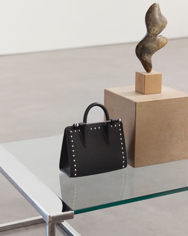 Studded black leather top-handle bag on a glass table, with a sculpture displayed behind it in a neutral, minimalist setting.