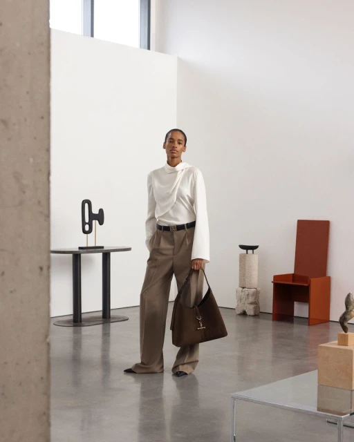 A woman in a white top and brown pants stands in a minimalist space, holding a brown shoulder bag, surrounded by modern art objects.