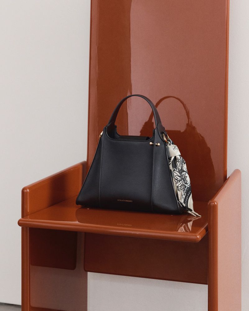 Black leather handbag on a shiny brown chair with a white background, featuring a decorative scarf accent for fashion accessory appeal.