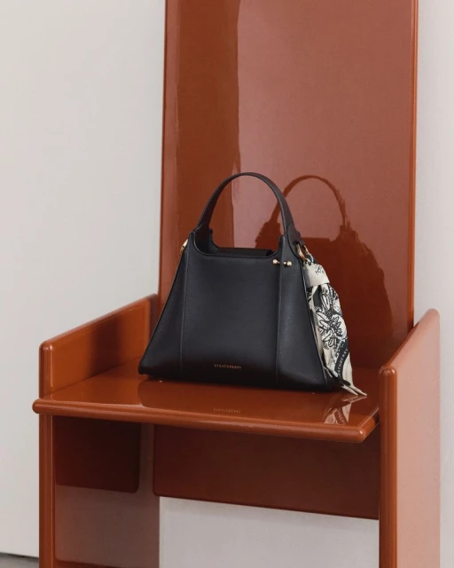 Black leather handbag on a shiny brown chair with a white background, featuring a decorative scarf accent for fashion accessory appeal.