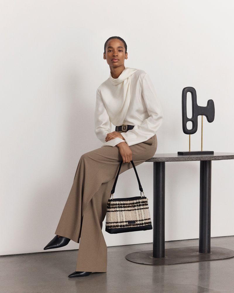 Elegant woman in neutral tones posing seated with a patterned handbag indoors against a neutral background, fashion photography.