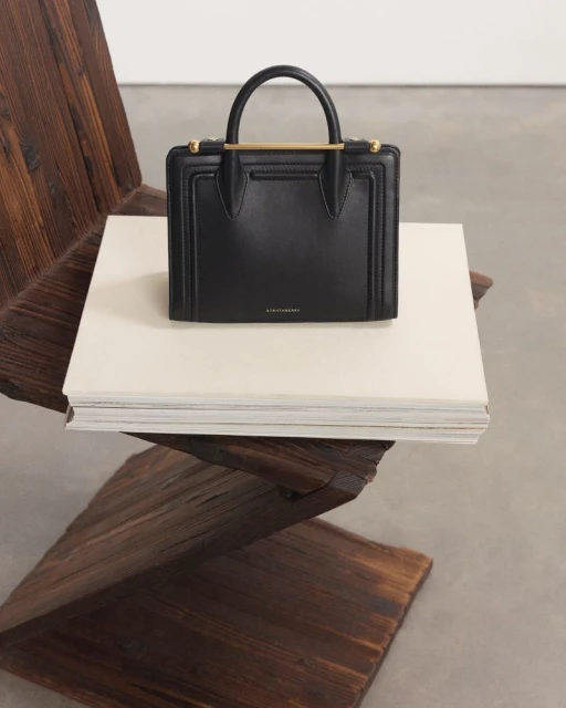 Studio shot of a black leather handbag with a gold metal bar on a wooden chair, placed on top of a stack of white books.