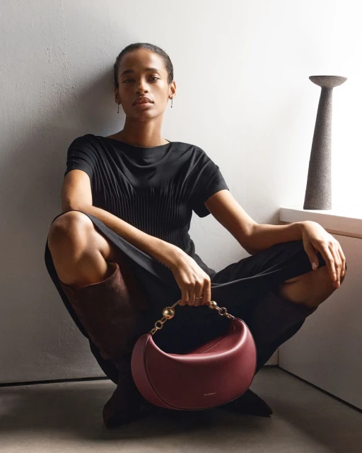 Woman poses indoors, seated and holding a burgundy purse with a gold chain. An abstract decorative vase is on a window sill.
