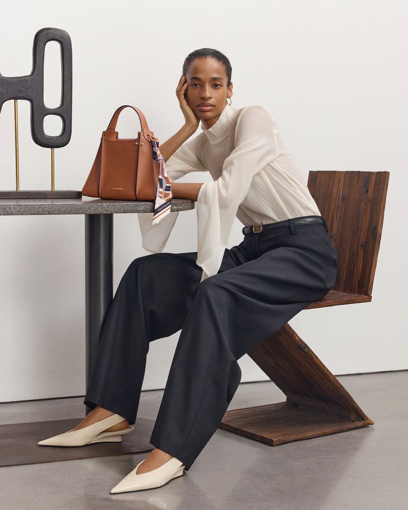 Adult woman models a brown leather purse while seated on a wooden chair. She wears a white top, dark pants, and light-colored shoes.