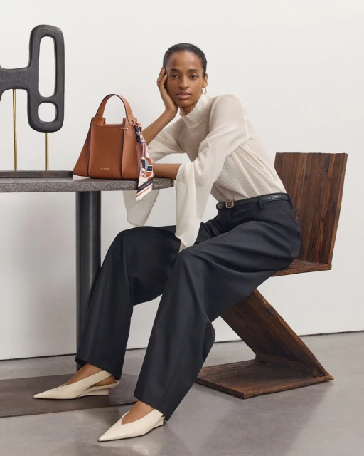 Adult woman models a brown leather purse while seated on a wooden chair. She wears a white top, dark pants, and light-colored shoes.