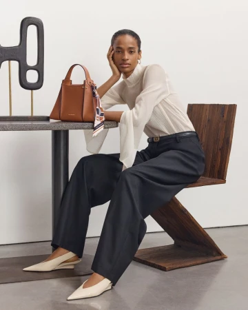 Adult woman models a brown leather purse while seated on a wooden chair. She wears a white top, dark pants, and light-colored shoes.