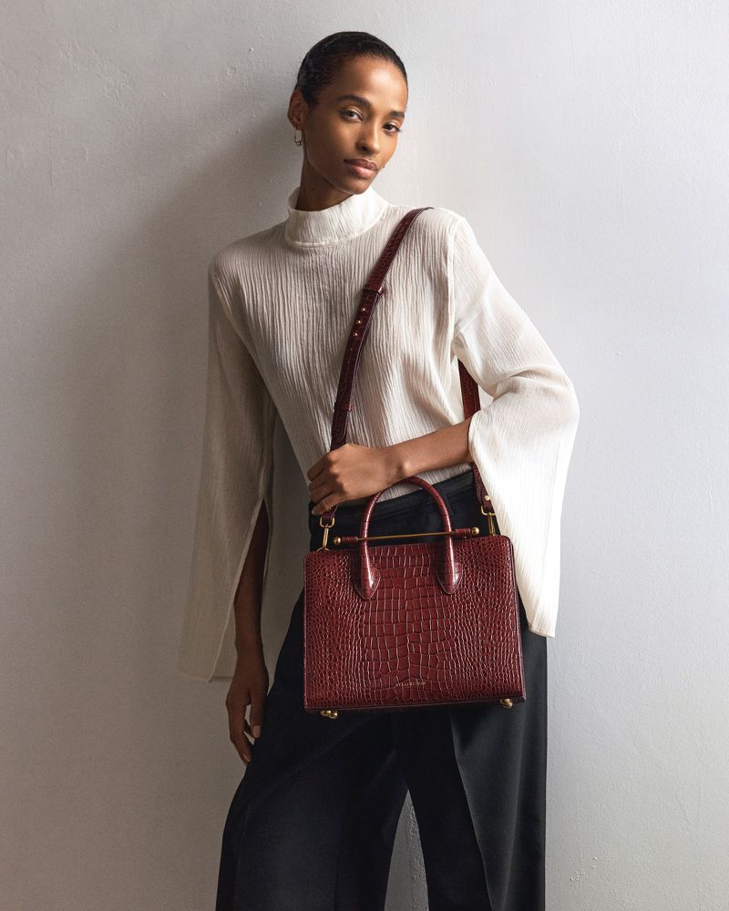 Elegant woman in a flowing white blouse and black trousers, holding a stylish red crocodile-patterned leather handbag with a shoulder strap.