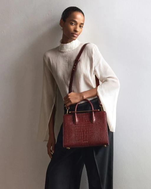Elegant woman in a flowing white blouse and black trousers, holding a stylish red crocodile-patterned leather handbag with a shoulder strap.