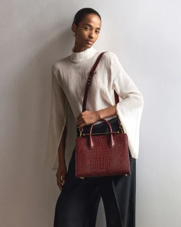 Elegant woman in a flowing white blouse and black trousers, holding a stylish red crocodile-patterned leather handbag with a shoulder strap.