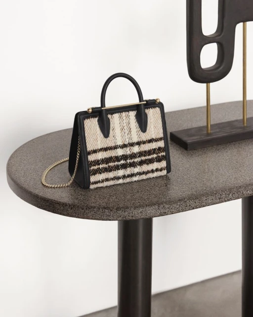 Striped fabric handbag with black trim and gold chain strap on a mottled grey table next to a dark brown abstract sculpture.
