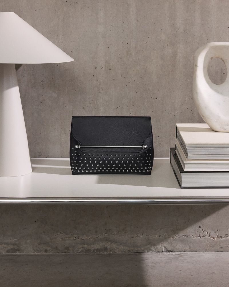 Black studded clutch purse on a white shelf alongside a modern lamp and art object, set against a concrete wall.