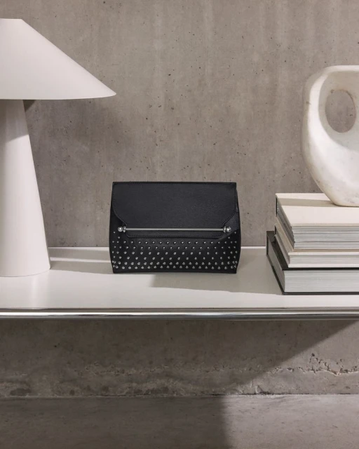 Black studded clutch purse on a white shelf alongside a modern lamp and art object, set against a concrete wall.