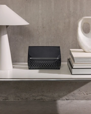 Black studded clutch purse on a white shelf alongside a modern lamp and art object, set against a concrete wall.