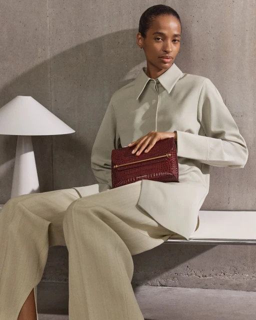 Woman in beige suit holds burgundy alligator clutch with gold hardware. Neutral backdrop and soft diffused lighting in studio setting.