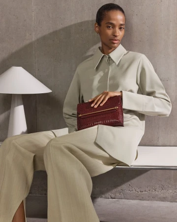 Woman in beige suit holds burgundy alligator clutch with gold hardware. Neutral backdrop and soft diffused lighting in studio setting.