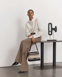 Model seated on a dark table holding a tweed handbag, wearing a white blouse, brown trousers and black heels against a plain white wall