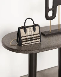 Structured tweed handbag with black leather trim and gold chain strap resting on a grey stone console table.