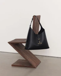 Black leather shoulder bag draped over a sculptural wooden zig-zag chair against a plain white wall and grey floor.