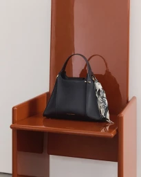 Black leather handbag with patterned silk scarf resting on a glossy orange chair against a light neutral background