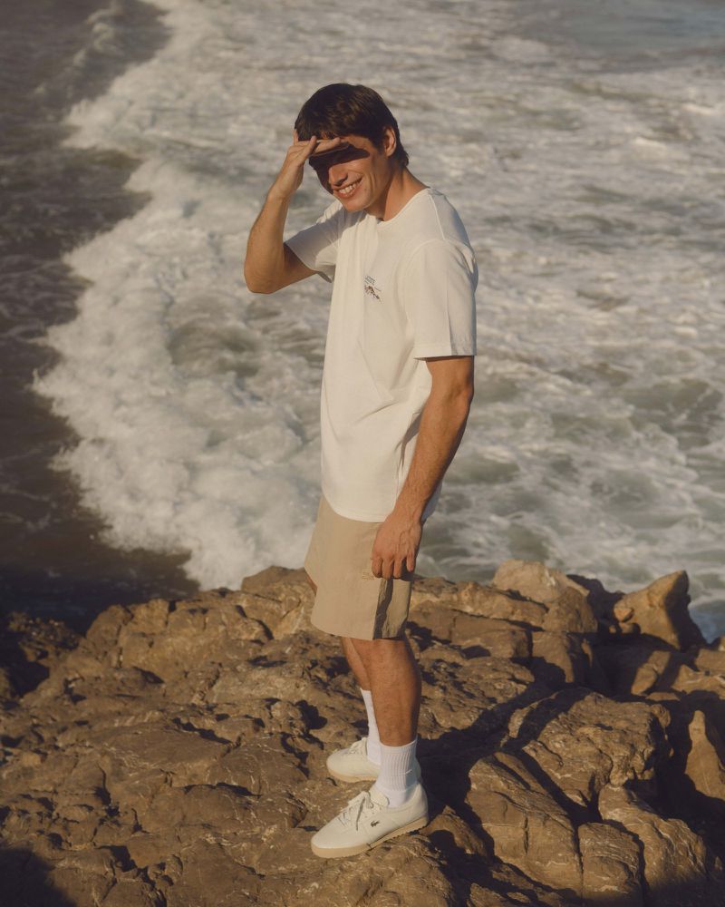 Man in t-shirt shields eyes on rocky coast with crashing waves in background. Coastal fashion shot. Tanned skin, white sneakers, shorts.