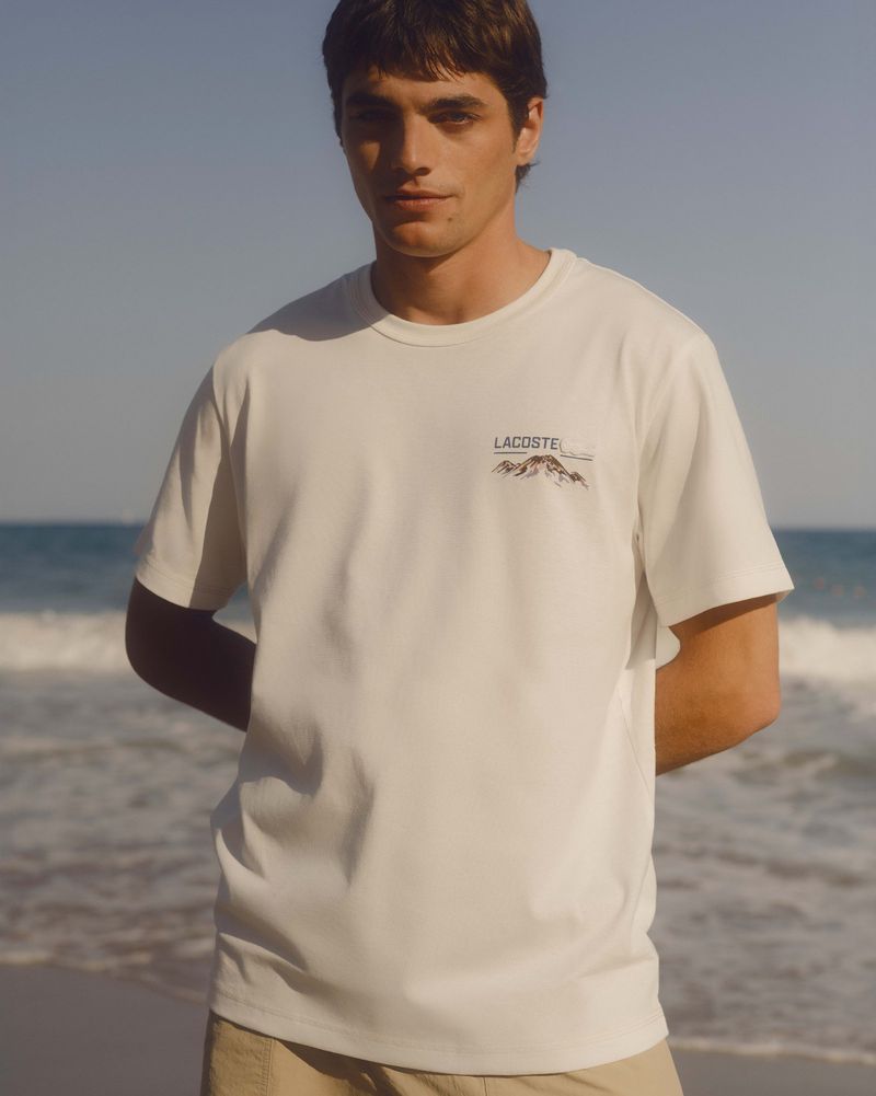 A young man poses wearing a white Lacoste T-shirt with a mountain graphic, in front of a beach and ocean backdrop.