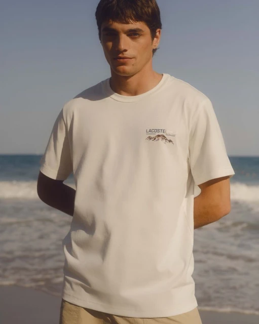 A young man poses wearing a white Lacoste T-shirt with a mountain graphic, in front of a beach and ocean backdrop.