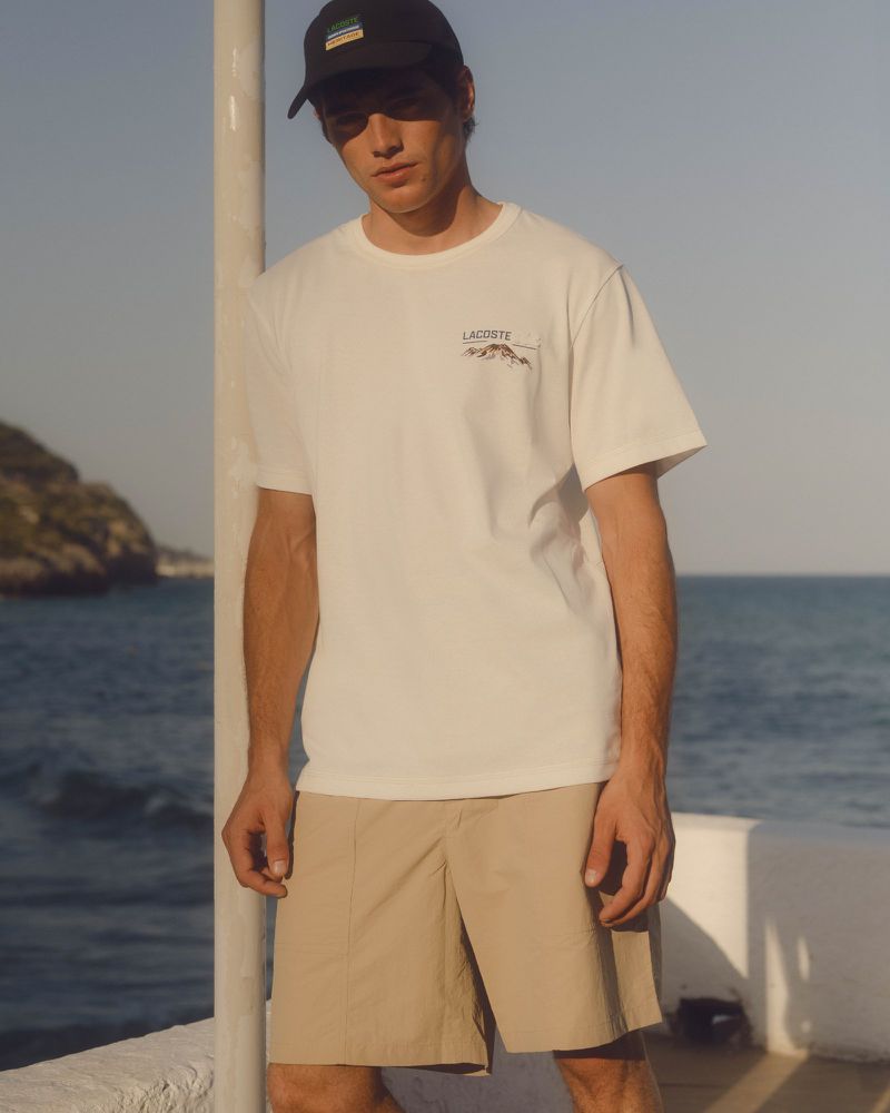 A young adult male poses in a white t-shirt and khaki shorts near the ocean, wearing a baseball cap and standing by a white pillar.
