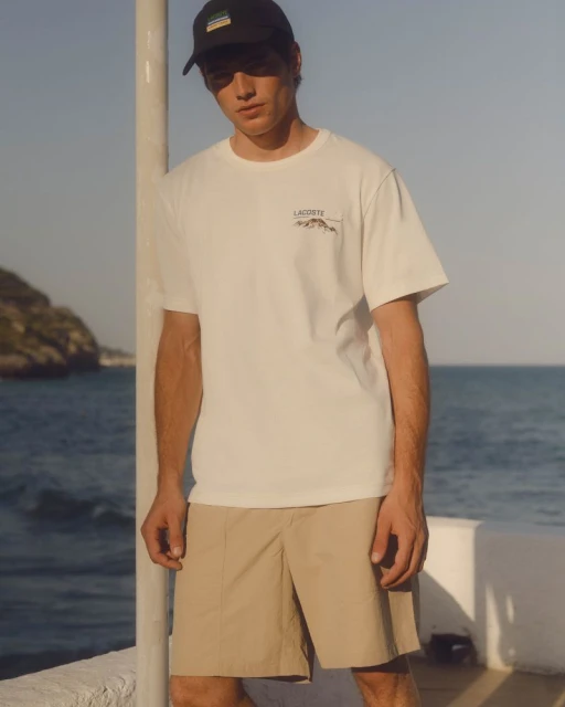 A young adult male poses in a white t-shirt and khaki shorts near the ocean, wearing a baseball cap and standing by a white pillar.