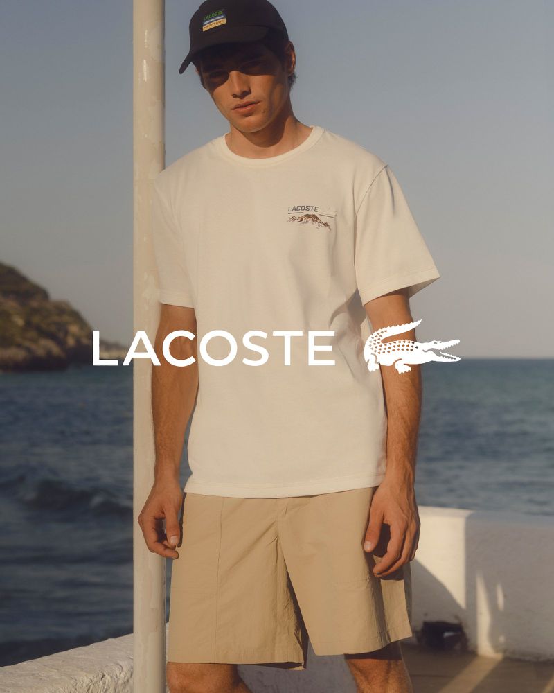 A young adult male poses outdoors wearing a white Lacoste T-shirt, khaki shorts, and a black baseball cap, in a fashion editorial shot.