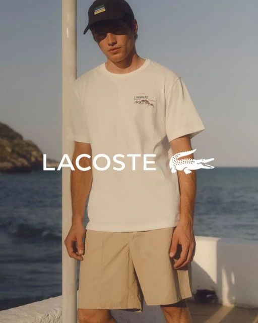 A young adult male poses outdoors wearing a white Lacoste T-shirt, khaki shorts, and a black baseball cap, in a fashion editorial shot.