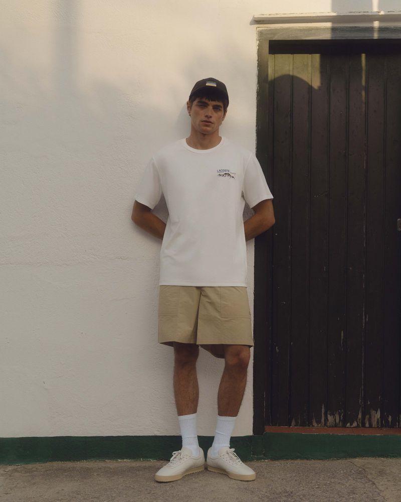 Man in casual attire stands against white wall with black door, wearing white t-shirt, khaki shorts, socks, sneakers and a baseball cap.