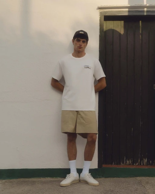 Man in casual attire stands against white wall with black door, wearing white t-shirt, khaki shorts, socks, sneakers and a baseball cap.