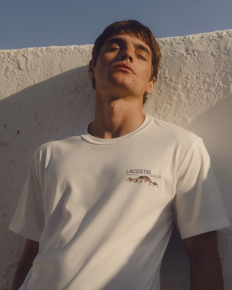 Young man in a white Lacoste t-shirt stands against a textured white wall in soft sunlight, showcasing a casual, summery fashion style.