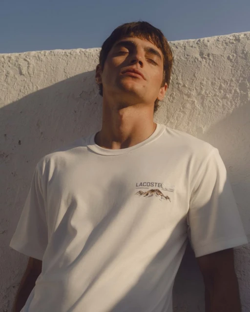 Young man in a white Lacoste t-shirt stands against a textured white wall in soft sunlight, showcasing a casual, summery fashion style.