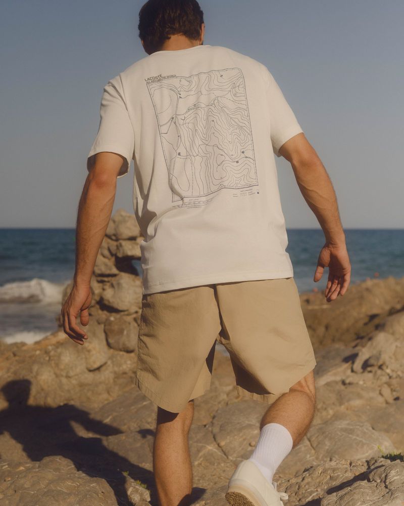 Adult male wearing a white t-shirt and khaki shorts walks on rocky terrain by the ocean on a sunny day with a clear blue sky.