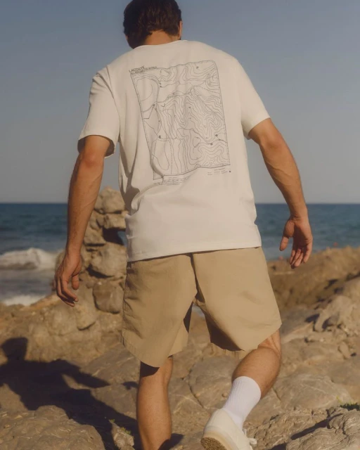 Adult male wearing a white t-shirt and khaki shorts walks on rocky terrain by the ocean on a sunny day with a clear blue sky.