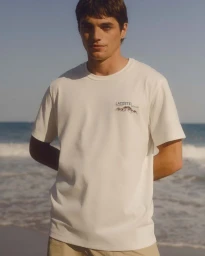 A young man wearing a white t-shirt standing on a sandy beach with the ocean in the background.