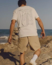Man from behind wearing a white t-shirt and tan shorts walking near a rocky coast by the ocean.