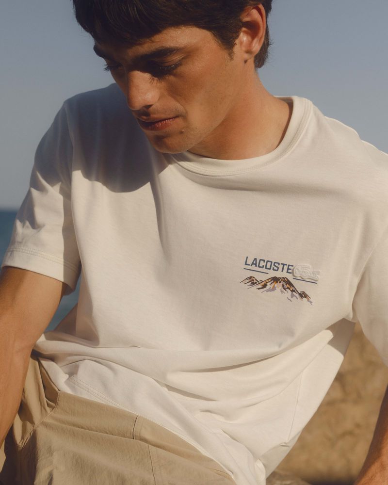 Close-up portrait of a man wearing a white Lacoste t-shirt with graphic print and tan shorts under bright natural daylight