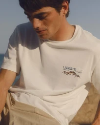 Close-up portrait of a man wearing a white Lacoste t-shirt with graphic print and tan shorts under bright natural daylight