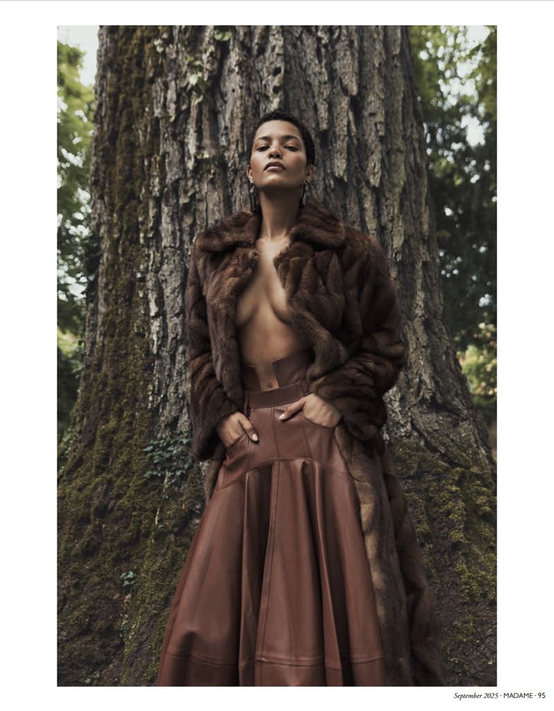 Woman in fur coat and leather skirt poses in front of a large tree in a natural outdoor setting during daytime.