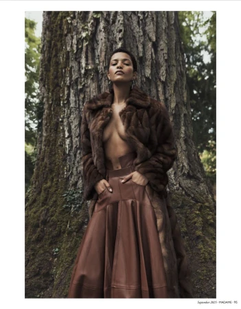Woman in fur coat and leather skirt poses in front of a large tree in a natural outdoor setting during daytime.