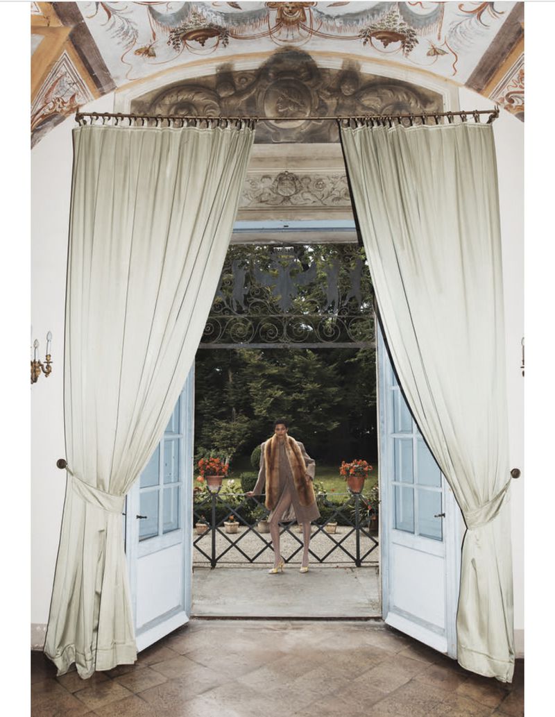 Fashion shot of an adult model in a tan coat with fur collar standing on a balcony viewed through open French doors with curtains.