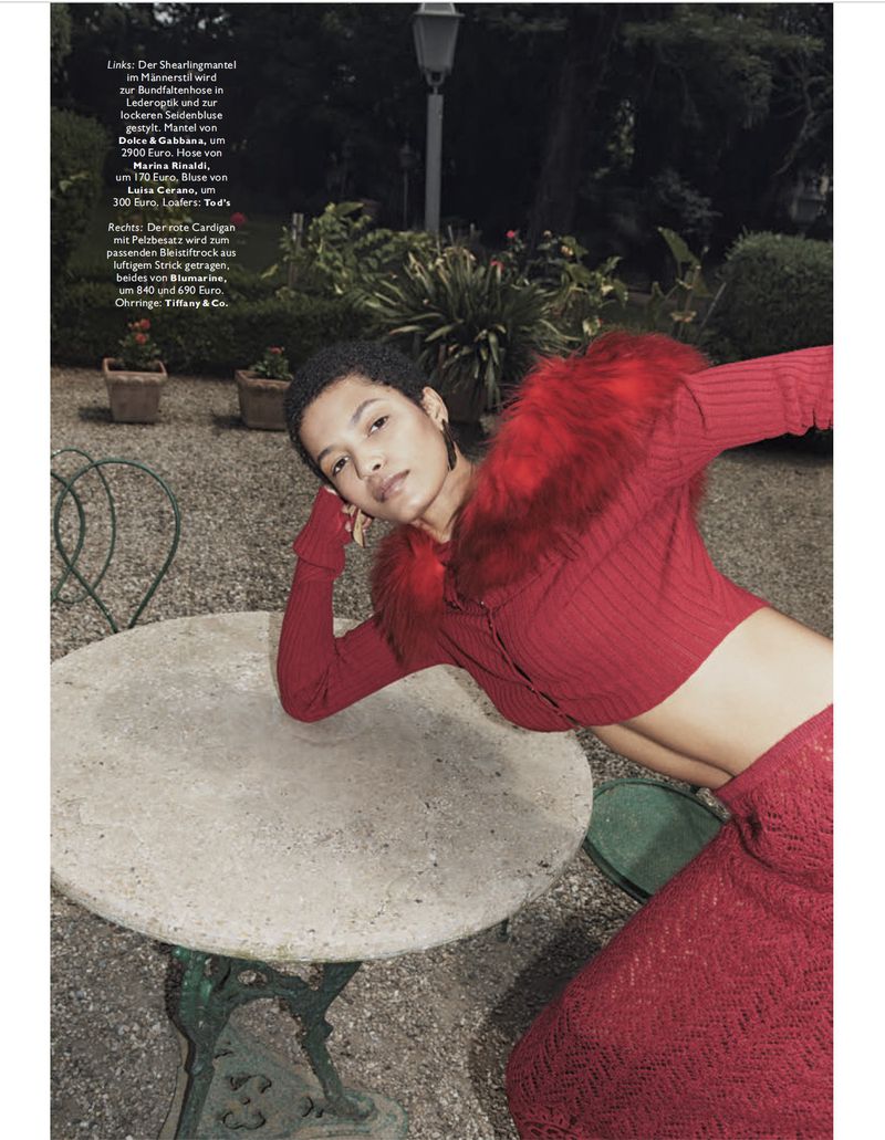 A woman with short hair in a red cardigan with fur trim and a red skirt is lying on an outdoor table in a garden setting.